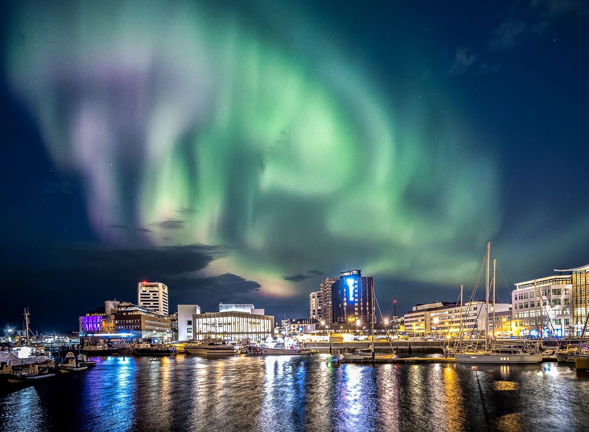 Byhavn om natten med nordlys i grønt og lilla over bygninger og båter