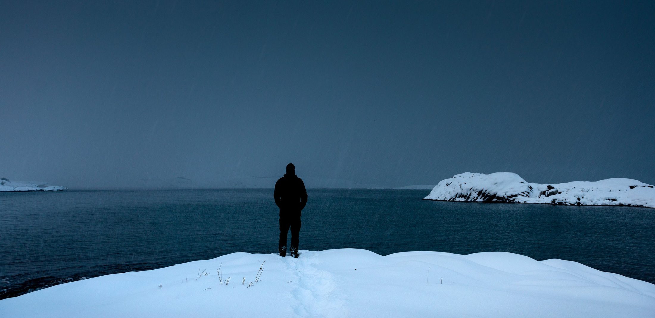 En person står på en snødekt kolle og ser ut over havet