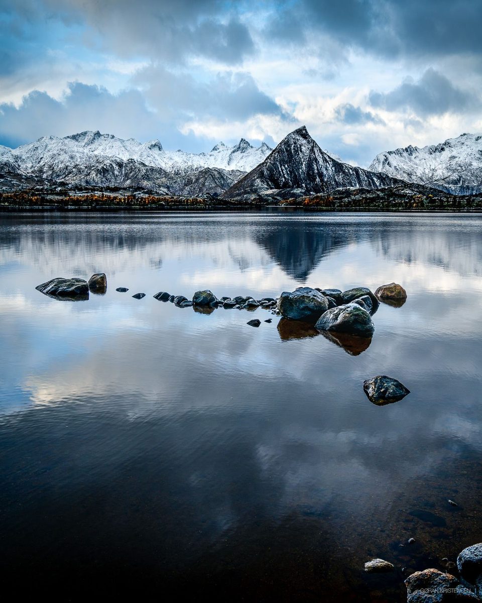 Fjell speilet i stille vann, med en rekke steiner i vannflaten og snø på toppene