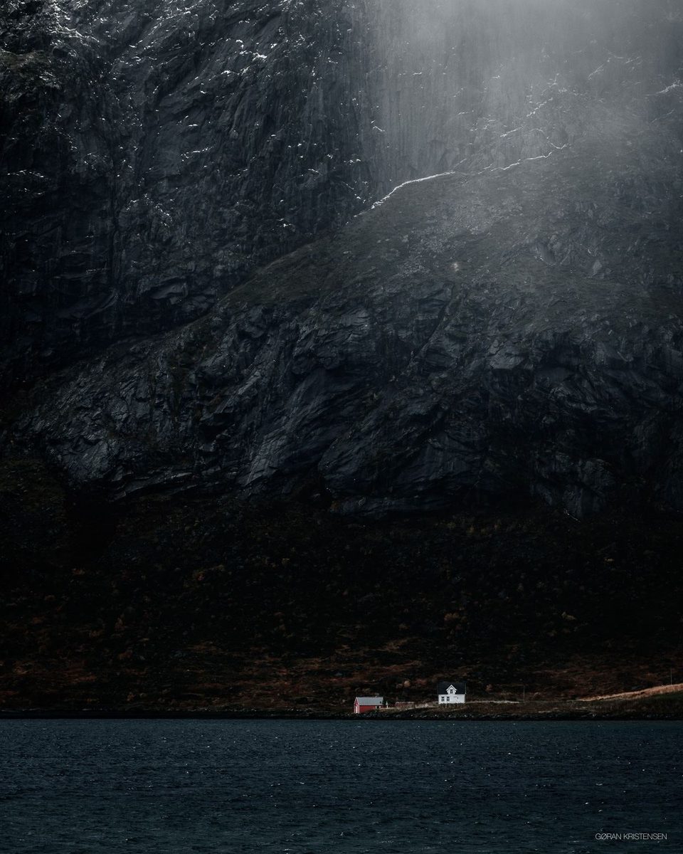 Mørk fjellside med et lysskjær som bryter gjennom, og et lite rødt/hvitt hus ved vannkanten