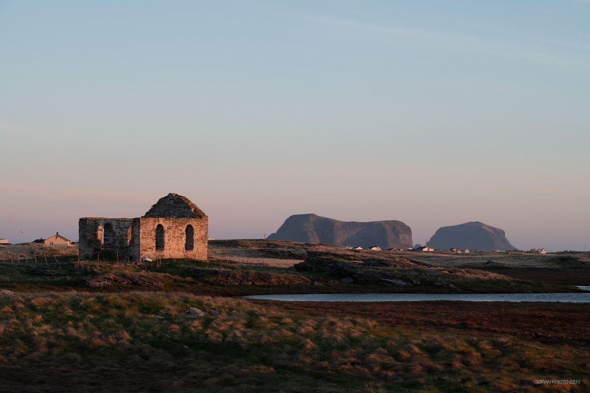 Ruinene av en gammel steinkirke i kveldslys, med to markerte fjelltopper i havet i bakgrunnen