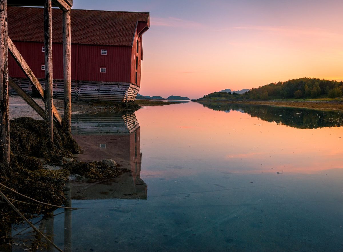 Rødt sjøhus på stylter ved vannkanten, speilet i stille vann ved solnedgang