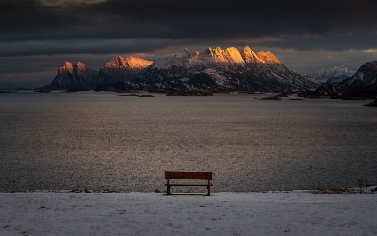 Tom benk med utsikt over en fjord og dramatisk solnedgangslys på fjell