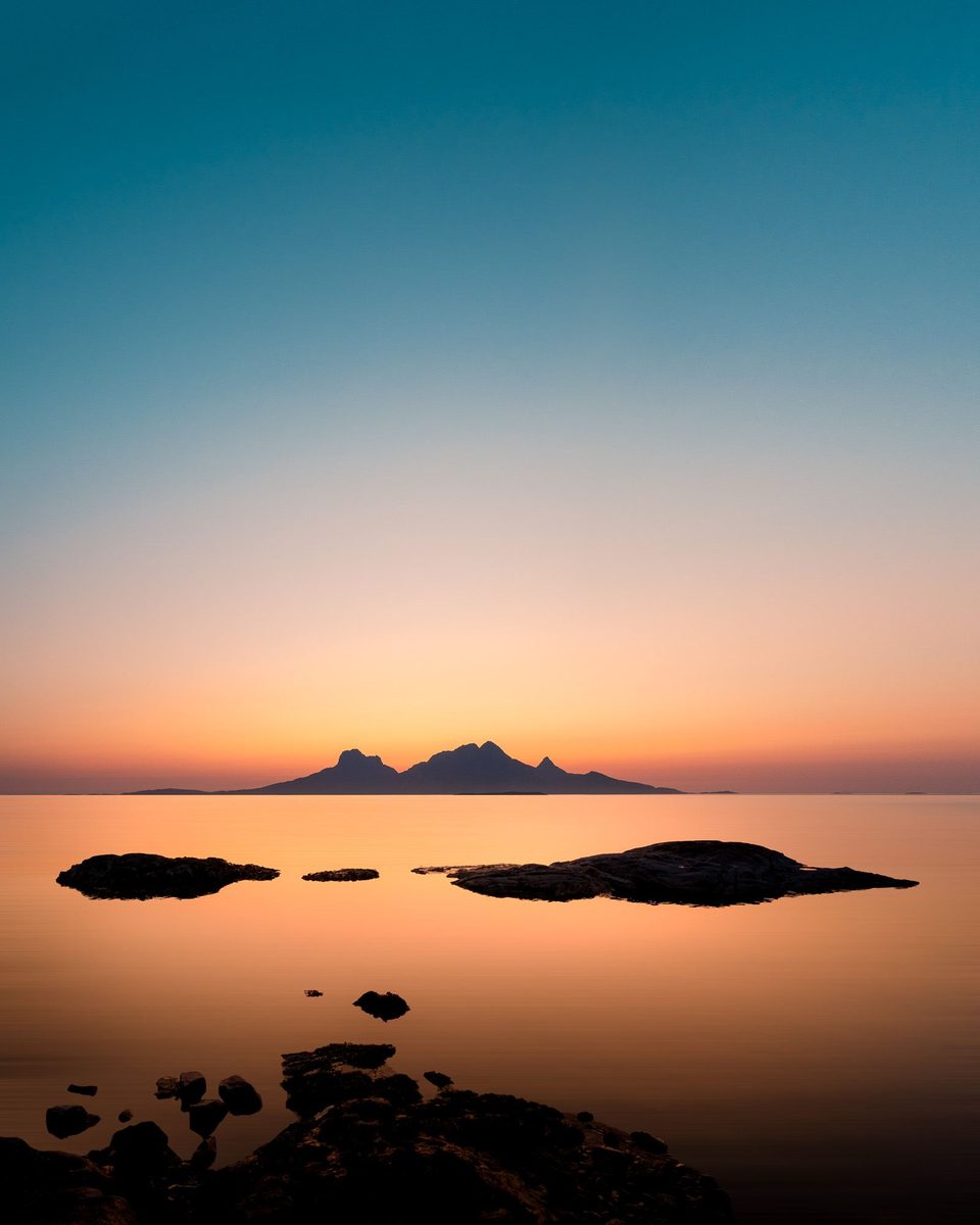 Fjell-silhuett på en øy i horisonten, med skjær som stikker opp av stille, fargerikt vann ved solnedgang